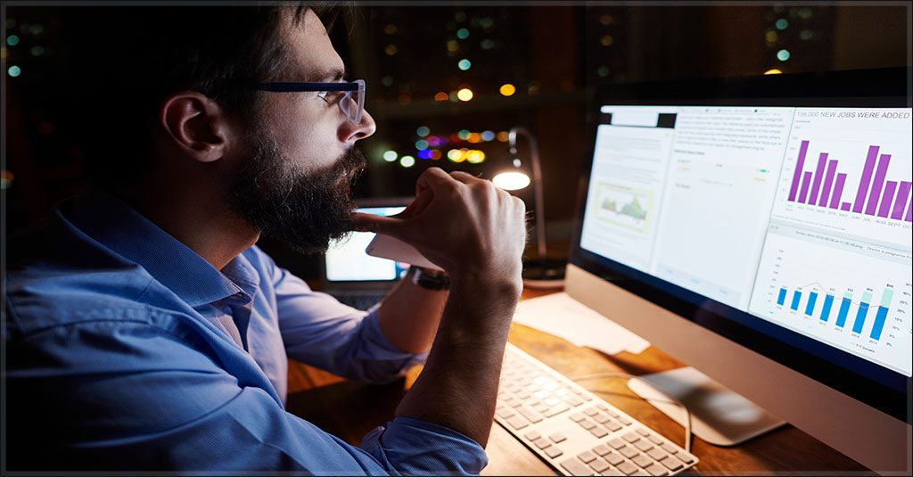 a young, bearded white man in a dress shirt looks pensively at charts on a computer monitor